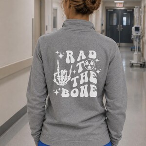 May include: A heather gray quarter-zip sweatshirt featuring a white graphic on the back. The design includes a skeleton hand making a rock-and-roll gesture, a radiation symbol, and the text "RAD TO THE BONE" in a retro, wavy font with stars.