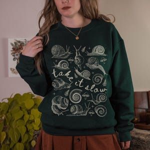 Puede incluir: Sudadera verde oscuro con un gr&aacute;fico de caracoles y setas color crema. Las palabras "take it slow" est&aacute;n escritas en cursiva. El dise&ntilde;o presenta varias ilustraciones de caracoles.