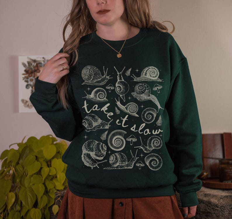 Puede incluir: Sudadera verde oscuro con un gr&aacute;fico de caracoles y setas color crema. Las palabras "take it slow" est&aacute;n escritas en cursiva. El dise&ntilde;o presenta varias ilustraciones de caracoles.