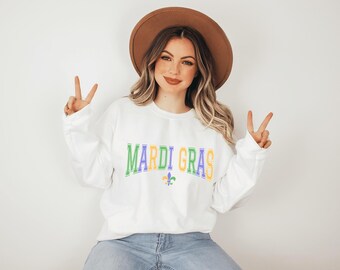Puede incluir: Sudadera blanca con el texto "Mardi Gras" en letras verdes, amarillas y moradas con un símbolo de flor de lis. La modelo lleva un sombrero marrón y unos vaqueros azules.