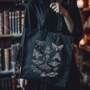 May include: Black tote bag featuring a collection of detailed moth illustrations in a cream color. The moths are arranged in a grid pattern with small star accents. The bag is held by a person in a dark-colored outfit, with a bookshelf in the background.