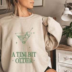 May include: A beige sweatshirt with a green graphic of a martini glass with olives and the text "A TINI BIT OLDER".