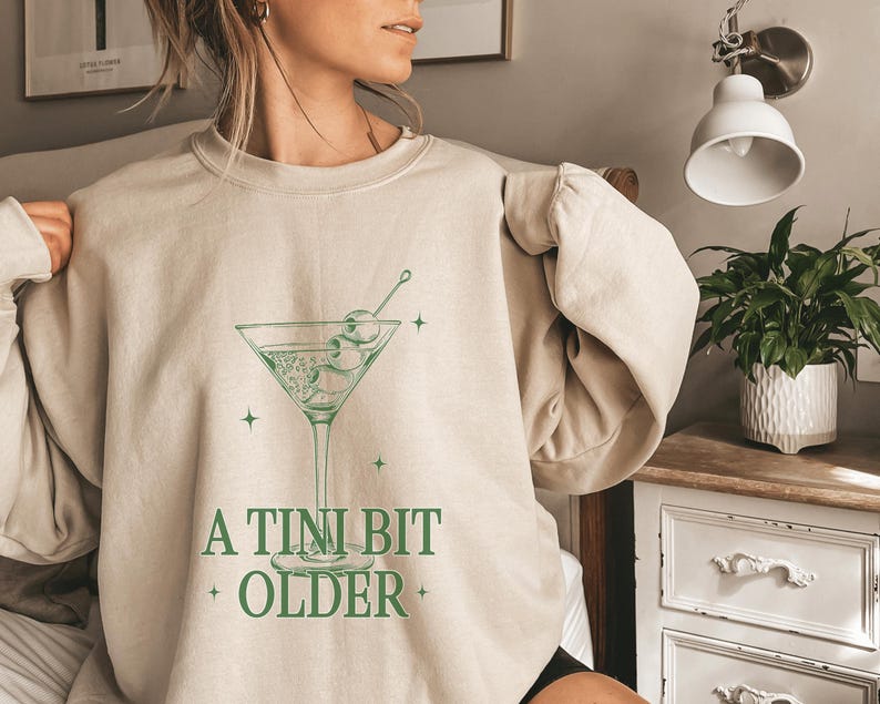 May include: A beige sweatshirt with a green graphic of a martini glass with olives and the text "A TINI BIT OLDER".