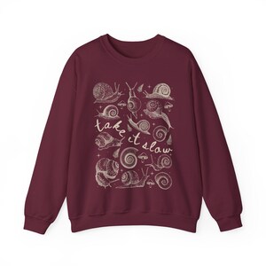 Puede incluir: Sudadera burdeos con una ilustraci&oacute;n crema de varios caracoles y setas. La frase "take it slow" est&aacute; escrita en cursiva. El dise&ntilde;o est&aacute; centrado en la parte delantera del cuello redondo de manga larga.