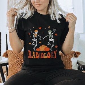 Puede incluir: Camiseta negra con un gráfico de dos esqueletos bailando con calabazas y el texto "RADIOLOGY".