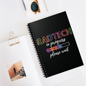 Könnte beinhalten: Ein schwarzes Notizbuch mit Spiralbindung, das den Text RADTECH in bunten Blumenbuchstaben zeigt, gefolgt von dem weißen Text in progress und please wait, mit einer Ladebalkengrafik mit kleinen bunten Blumen zwischen den Sätzen.