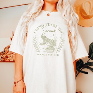 May include: Cream-colored t-shirt featuring a green frog illustration within a circular design. The text "FRESH FROM THE SWAMP" and "LOCALLY SOURCED" are printed above and below the frog graphic. The shirt has short sleeves and a crew neck.