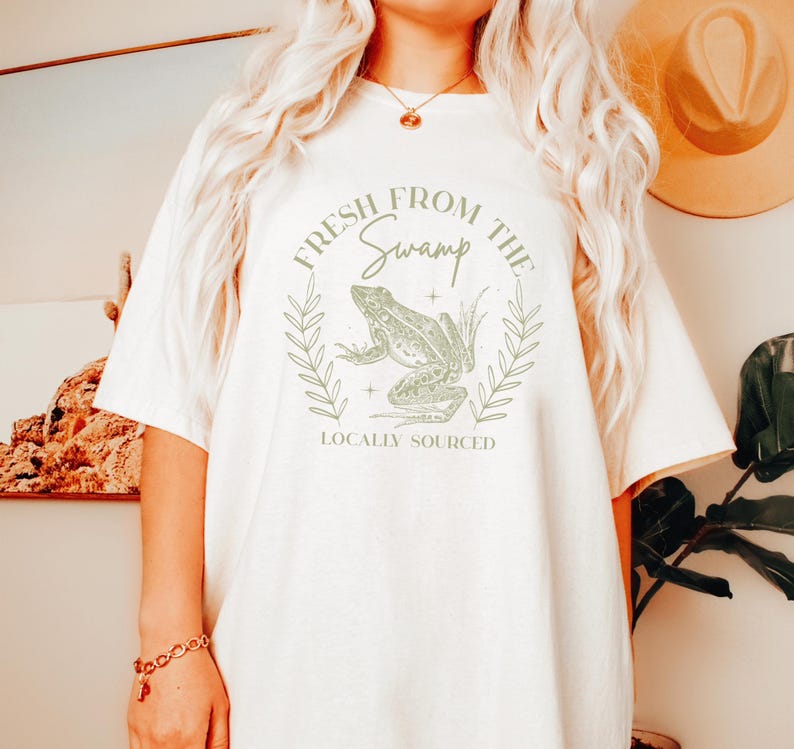 May include: Cream-colored t-shirt featuring a green frog illustration within a circular design. The text "FRESH FROM THE SWAMP" and "LOCALLY SOURCED" are printed above and below the frog graphic. The shirt has short sleeves and a crew neck.