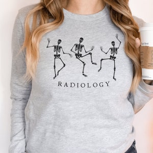 Op de afbeelding: Grijs longsleeve sweatshirt met een afbeelding van drie dansende skeletten en de tekst "RADIOLOGY".