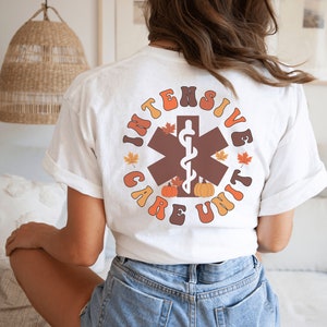 Peut inclure: T-shirt blanc avec un motif r&eacute;tro au dos. Le motif pr&eacute;sente les mots "Intensive Care Unit" en orange et marron, entourant un symbole m&eacute;dical marron avec un serpent blanc. Des feuilles d'automne et des citrouilles compl&egrave;tent le design.