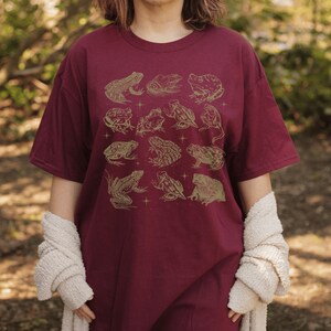 Pode incluir: Uma camiseta vinho com um padr&atilde;o de sapos dourados. Os sapos est&atilde;o em v&aacute;rias poses, alguns est&atilde;o sentados, outros est&atilde;o pulando e outros est&atilde;o nadando. A camiseta tem um caimento solto e um decote redondo.