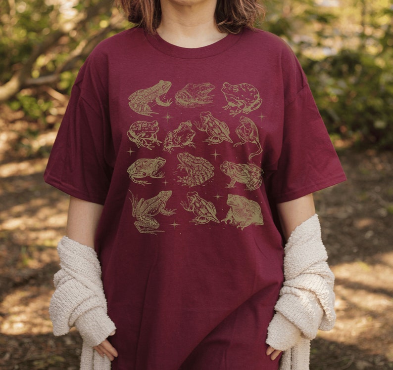 Pode incluir: Uma camiseta vinho com um padr&atilde;o de sapos dourados. Os sapos est&atilde;o em v&aacute;rias poses, alguns est&atilde;o sentados, outros est&atilde;o pulando e outros est&atilde;o nadando. A camiseta tem um caimento solto e um decote redondo.