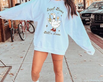 Puede incluir: Sudadera blanca con un dibujo de un ganso sosteniendo una botella y una copa de vino. El texto dice "Legal Goose Gett'n Loose". El ganso lleva zapatos rojos. Estilo casual.