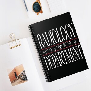 May include: A black spiral-bound notebook with the words "RADIOLOGY DEPARTMENT" in white, with a red line and dancing skeleton figures. The notebook is open on a white surface with other items.
