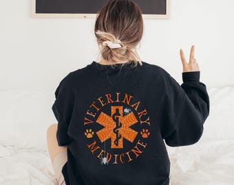 May include: Black sweatshirt with a white circle graphic on the back. The graphic features a red medical symbol with a snake and spider web design. The text "Veterinary Medicine" is written around the symbol.