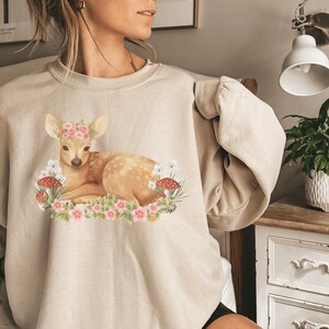 May include: Beige sweatshirt featuring a fawn with a floral crown, surrounded by flowers and mushrooms. The fawn is lying down, with a light brown coat and white spots. The design is a whimsical illustration.