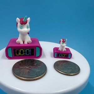 May include: Two pink digital alarm clocks with unicorn figurines on top. The larger clock displays "00:01" and the smaller clock displays "10:00". Two coins are in the foreground. The clocks are on a white surface with a blue background.