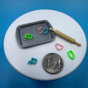 Könnte beinhalten: Miniatur-Backset mit einem grauen Tablett, bunten Ausstechern und einem kleinen Nudelholz. Die Ausstecher sind in Form eines Hasen, eines Eies, einer Karotte und einer Blume. Eine Münze ist zum Maßstab enthalten.