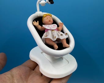 以下が含まれることがあります： 金色の星柄の白いドレスと花柄のよだれかけを身につけたブロンドのミニチュアドールが、白いプラスチック製のベビーバウンサーに乗っています。バウンサーには、黒と白の吊り下げ式おもちゃが付いた小さなモビールが取り付けられています。