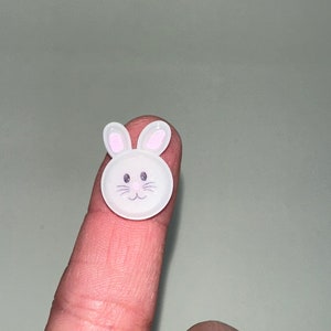 以下が含まれることがあります： ウサギの顔が付いた白いプラスチックボタン。ウサギはピンクの耳、黒い目、黒い鼻、黒い口をしています。
