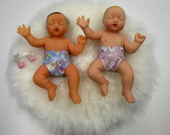 May include: Two miniature baby dolls with closed eyes and outstretched arms. One doll has a darker skin tone and wears a blue diaper with a shark print, while the other has a lighter skin tone and wears a pink diaper with a cat print.