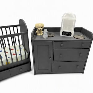 May include: A miniature dark grey wooden cot with a patterned mattress and a matching dark grey chest of drawers featuring three drawers and a cupboard door. On the chest sits a small beige crochet puppy, a clear plastic baby bottle, and a white steriliser.