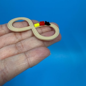 以下が含まれることがあります： ベージュ色の無限大の形をしたおもちゃの線路で、黄色、赤、黒の3つの小さな電車のおもちゃが付いています。青い背景に、手で線路が持たれています。おもちゃは、おそらくミニチュアモデルまたは装飾品です。