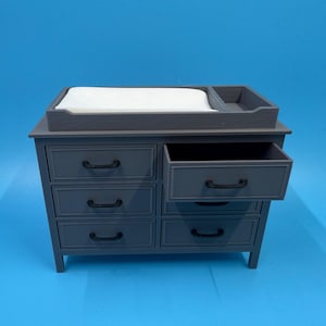 May include: A gray changing table with six drawers and a changing pad. The top of the table has a raised edge and a compartment. One drawer is open, revealing the interior. The handles are black.