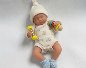 May include: A realistic baby doll wearing a cream-colored knit hat, a cream-colored onesie with a pattern of small brown bears and blue elephants, and light blue knit booties. The doll holds a yellow rattle in one hand and a multi-colored ring toy.