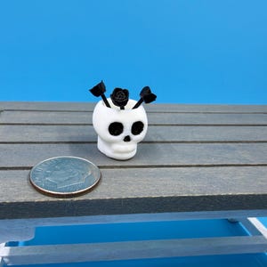 Könnte beinhalten: Ein kleiner, weißer, totenkopfförmiger Übertopf mit drei kleinen schwarzen Blumen, der neben einer Münze als Größenvergleich steht. Der Totenkopf hat schwarze Augenhöhlen und eine Nasenöffnung sowie eine glatte, abgerundete Oberfläche.