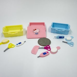 Op de afbeelding: Een verzameling miniatuur speelgoedaccessoires met drie kleine rechthoekige mandjes in geel, roze en blauw, met een Hello Kitty-afbeelding op het roze deksel. Bevat ook kleine kammen, scharen, thermometers en een muntstuk voor de schaal.