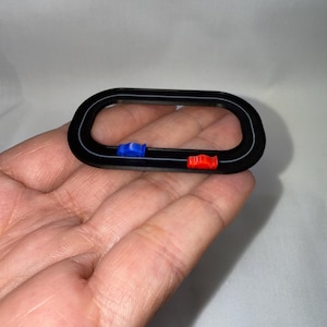 Könnte beinhalten: Eine kleine, schwarze ovale Rennstrecke mit einem blauen und roten Spielzeugauto. Die Rennstrecke hat einen weißen Streifen in der Mitte. Die Spielzeugautos sind klein und farbenfroh und verleihen dem Design eine spielerische Note.
