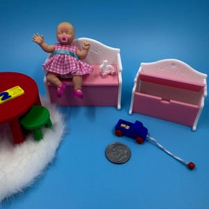 May include: A miniature dollhouse scene featuring a baby doll in a pink and white checked dress, seated on a pink bench. A blue toy car, a small white bunny, and a red table with a green stool are also present. A coin is included for scale.