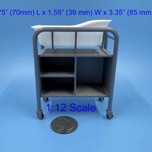 May include: A 1:12 scale miniature hospital bassinet with a grey frame and a white basin. The unit features three open storage compartments and four small wheels. Dimensions are 2.75 inches L x 1.55 inches W x 3.35 inches H, shown with a US quarter.