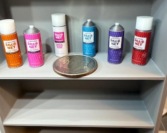 May include: Miniature Aqua Net hairspray cans in orange, pink, white, blue, purple, and red are arranged on a shelf. Each can has a white cap and the Aqua Net logo. A coin is placed in front of the cans. The shelf is part of a gray cabinet.