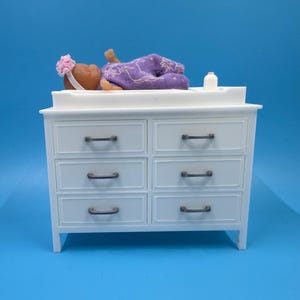 May include: A white changing table with six drawers and dark metal handles. A doll wearing a purple outfit and a pink headband lies on top of the table. A small white bottle is also on the table.