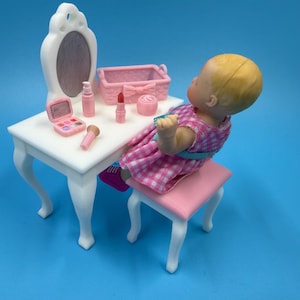 May include: A miniature white vanity table with an attached oval mirror, a matching white stool with a pink seat, and a doll wearing a pink and white checkered dress. Accessories include a pink basket, a makeup palette, a lipstick, and a brush.