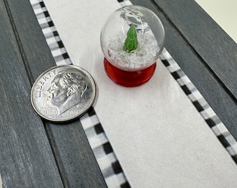 May include: A miniature snow globe with a small green Christmas tree inside. The globe is filled with white glitter and has a red base. A US quarter is shown for scale.