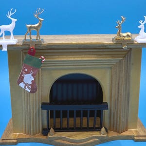 May include: A miniature wooden fireplace with a light brown finish, adorned with four decorative reindeer figurines in gold and white. A Christmas stocking with a reindeer design hangs from the mantel. The fireplace has a black grate and a blue background.