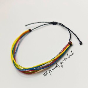 Puede incluir: Un brazalete trenzado de colores con hilos rojos, amarillos, azules, verdes y plateados. El brazalete está atado con un cordón negro y tiene cuentas de madera en los extremos. El texto "10% proceeds given back" está escrito sobre el fondo blanco.