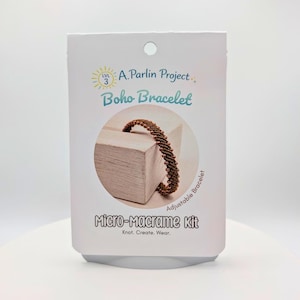 May include: A white rectangular packaging card for an A. Parlin Project Level 3 Boho Bracelet Micro-Macrame Kit. The card features text and a circular cutout showing a brown braided bracelet wrapped around a light-colored wooden block.