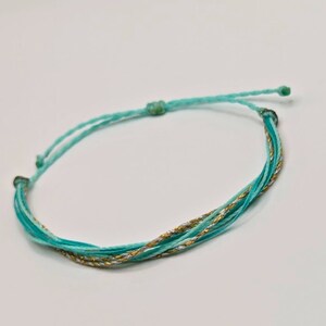 May include: A turquoise and gold friendship bracelet with a sliding knot closure.