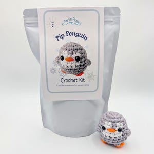 May include: A crochet kit for a Pip Penguin, featuring a gray and white penguin with an orange beak and feet. The kit includes a bag with a label that reads "Pip Penguin Crochet Kit" and "Crochet creations for sensory play."