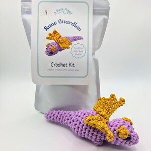 Puede incluir: Un kit de ganchillo para un peluche de Rune Guardian. El kit incluye un dragón morado con alas doradas. El embalaje incluye el texto "Rune Guardian Crochet Kit" y "Create a bean bag plushie".