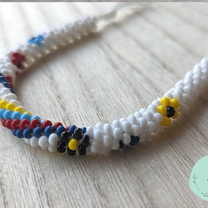 May include: White beaded bracelet with colorful flower accents. The bracelet is made with small white beads and features a repeating pattern of red, blue, yellow, and black beads. The flower accents are made with yellow and black beads.