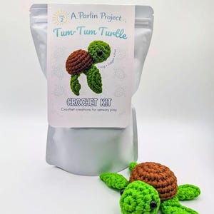 May include: A crochet kit for a Tum-Tum Turtle, with a finished turtle beside the package. The turtle has a brown shell and green head and limbs. The kit's package has the text "Tum-Tum Turtle" and "CROCHET KIT".