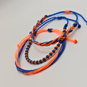 May include: Three friendship bracelets in blue and orange. One bracelet has blue beads, one is braided with orange and blue string, and the third is a solid orange string.