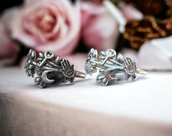 May include: Two silver rings with floral designs, each featuring a key, a hamsa hand, and a crescent moon. The rings are on a white surface with a blurred background of pink roses.