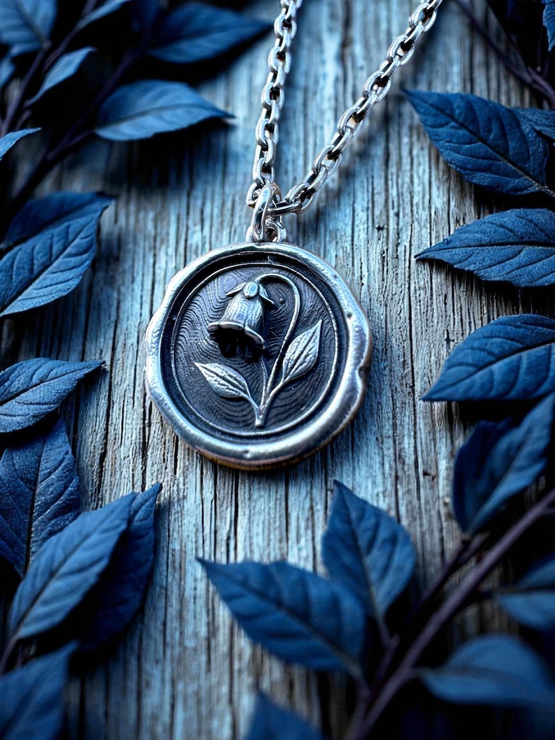 May include: A silver pendant necklace featuring a detailed floral design. The pendant, with a flower and leaves, hangs from a chain. The image is set against a wooden background with blue leaves, creating a nature-inspired aesthetic.