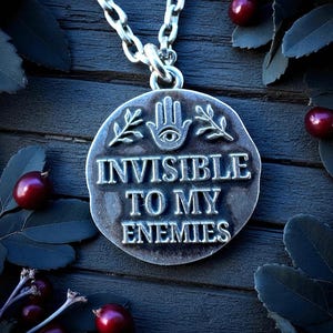 Sterling silver pendant engraved with the phrase ‘Invisible to My Enemies,’ featuring an all-seeing eye in a hand and leafy branches, shown on dark wood with berries and leaves